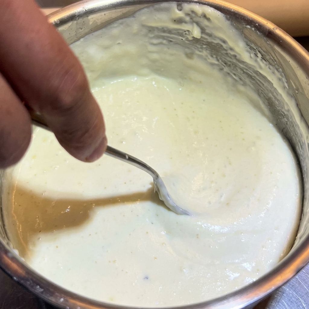 A metal spoon stirring a thick, creamy white cheese sauce in a stainless steel pot.