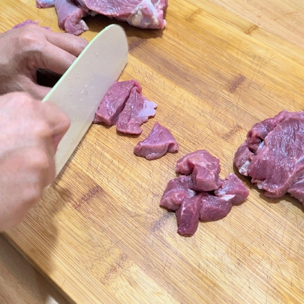 Hands slicing raw beef into small, uniform cubes on a wooden cutting board.