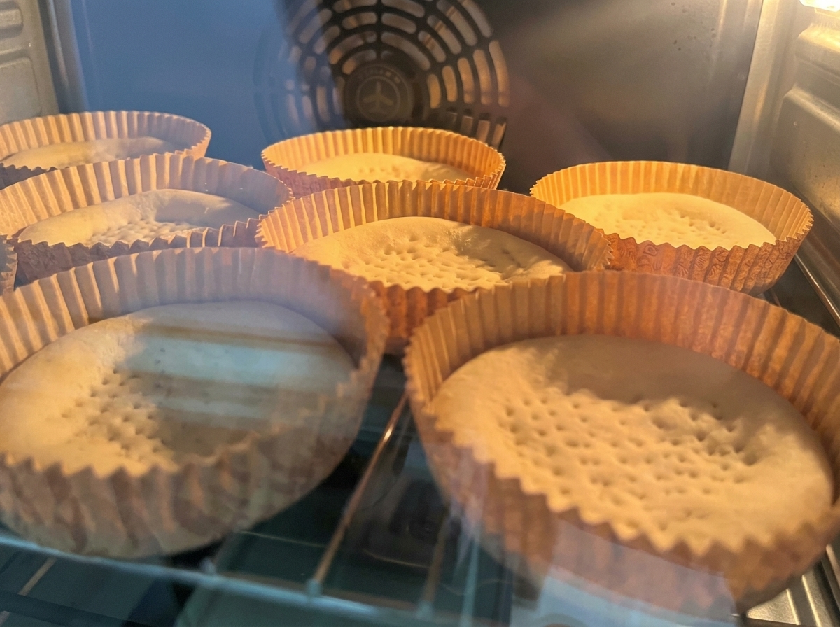 Empty pizza crusts baking in paper trays inside an illuminated oven.