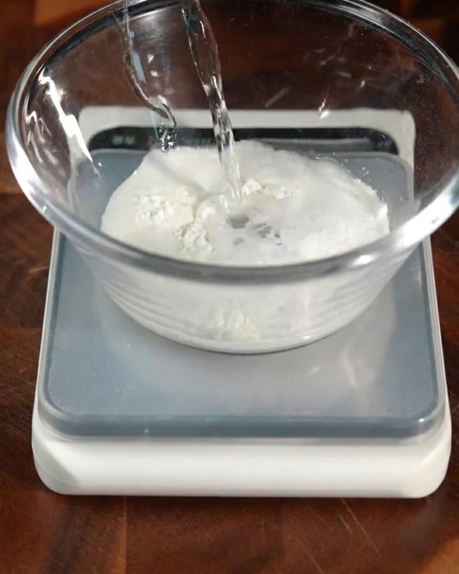 Warm yeast water being poured into a bowl containing flour and other dry ingredients.