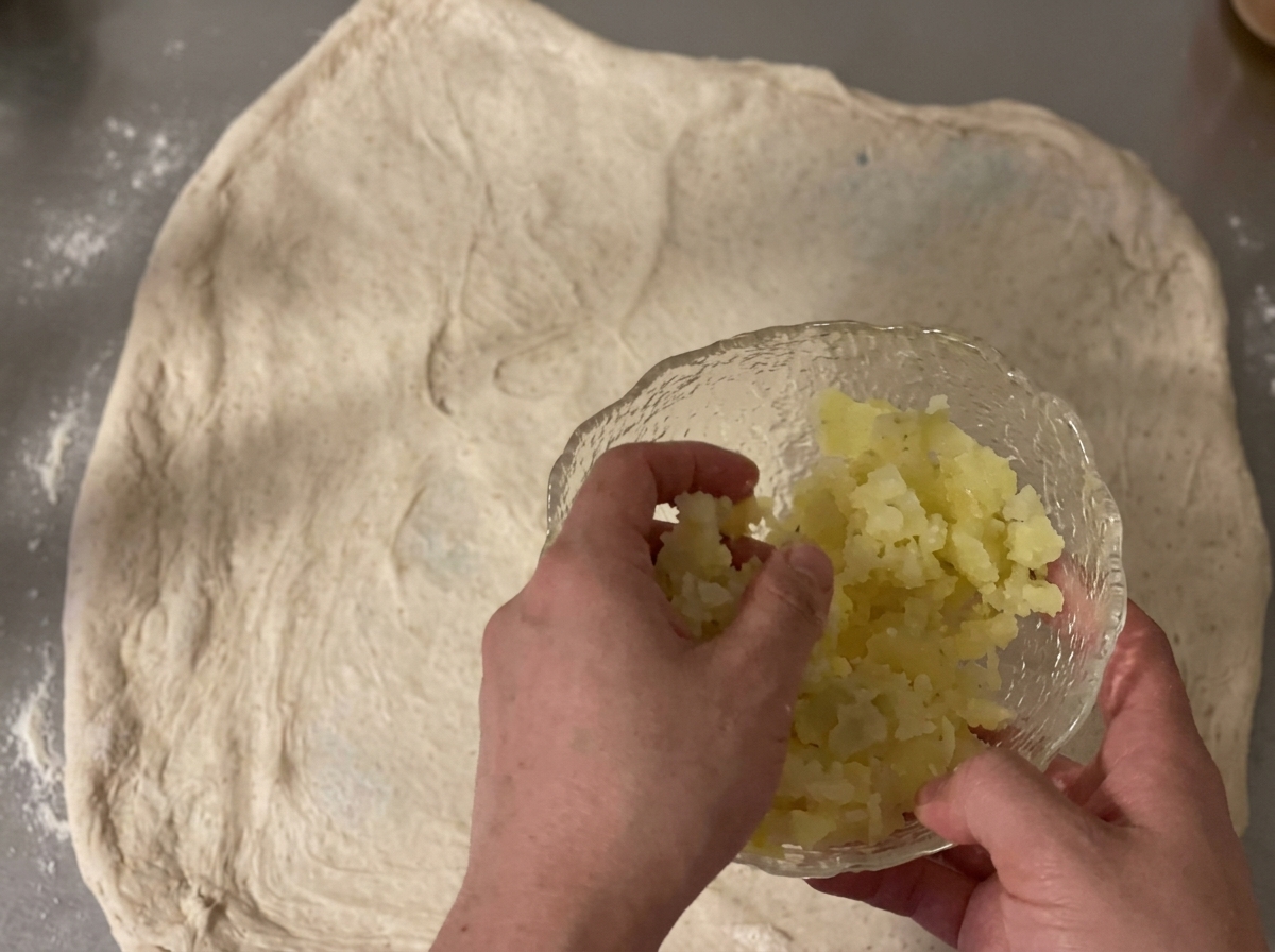 Diced potatoes and shredded cheddar cheese being scattered over a sheet of raw dough.