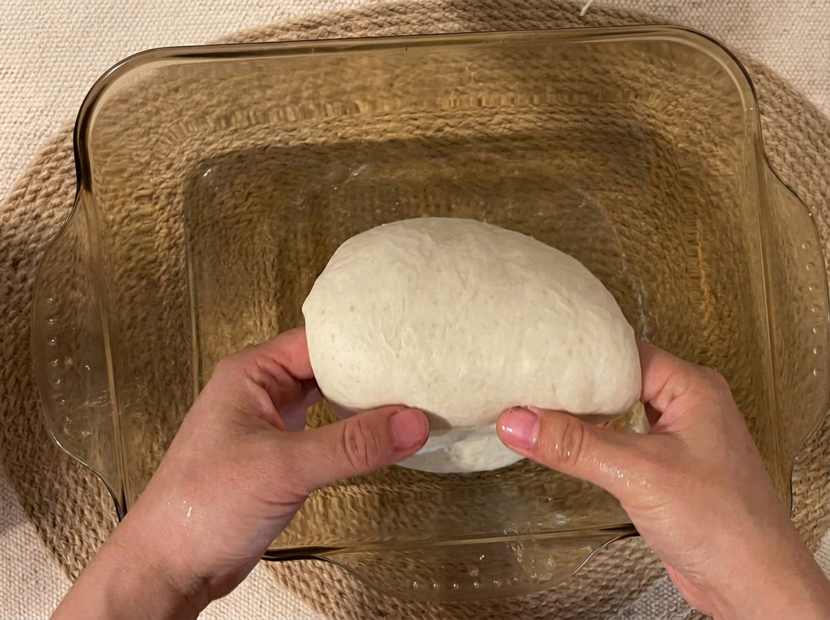 Dough being lifted from the center to initiate a coil fold inside a glass baking dish.
