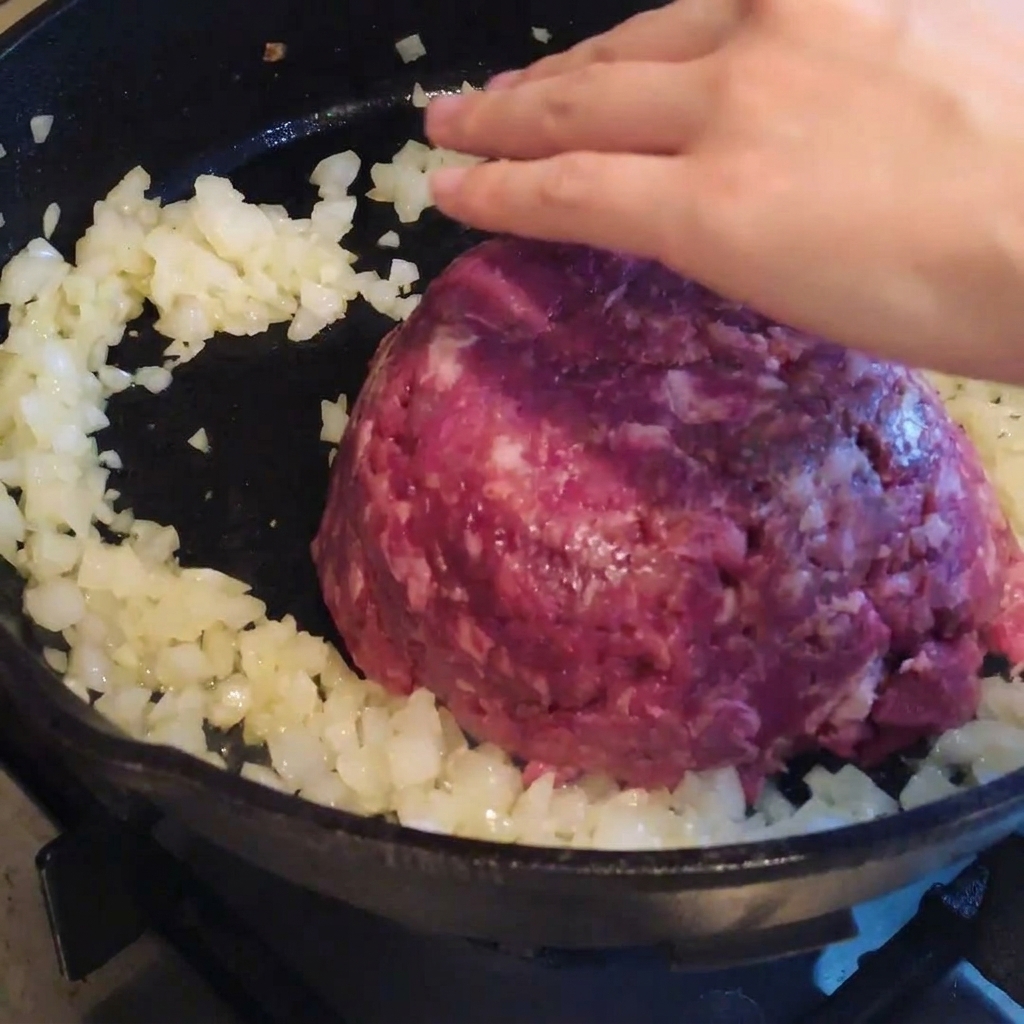 Ground beef being placed into a skillet with previously sautéed onions, ready to be stirred and seasoned.