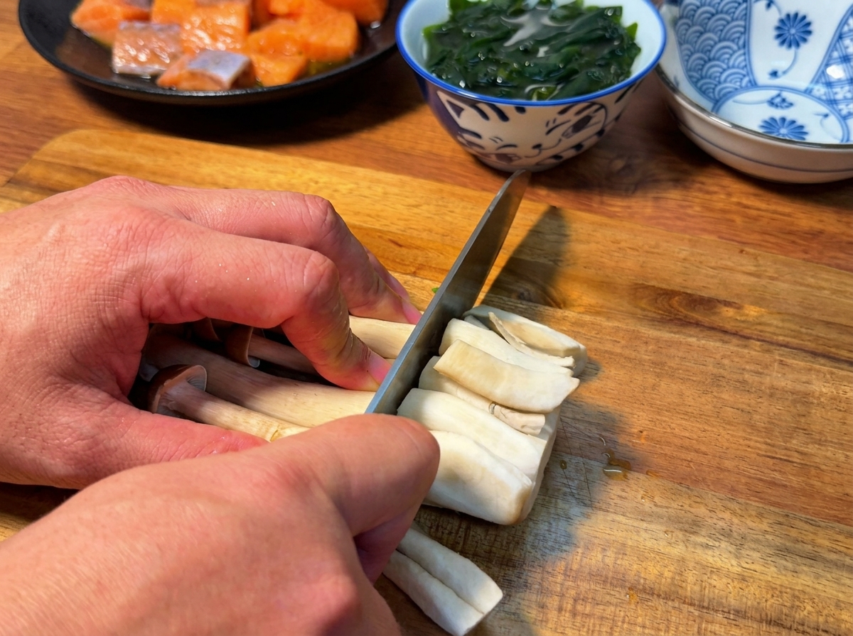 A close-up view of a knife cutting the roots off a bundle of shimeji mushrooms on a wooden cutting board.
