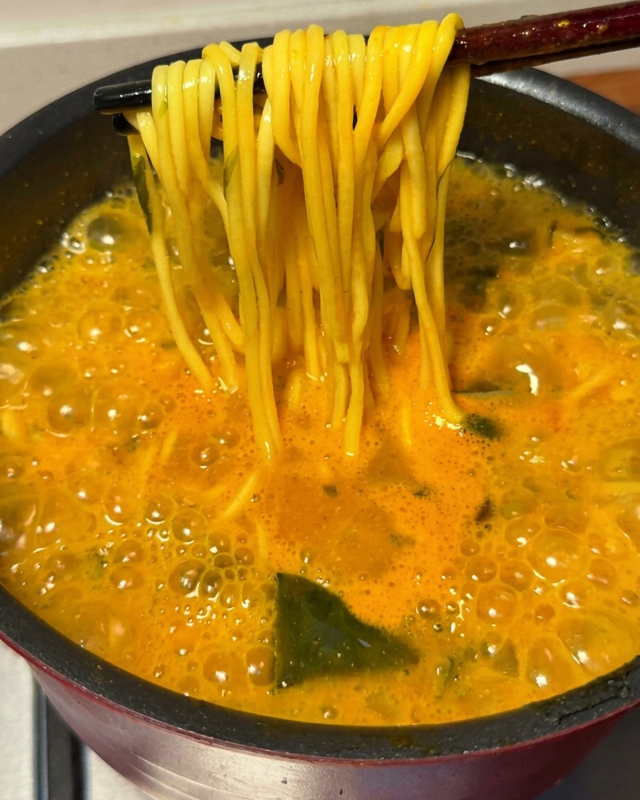 A pair of chopsticks lifting a bundle of golden cooked ramen noodles out of the bubbling spicy broth.