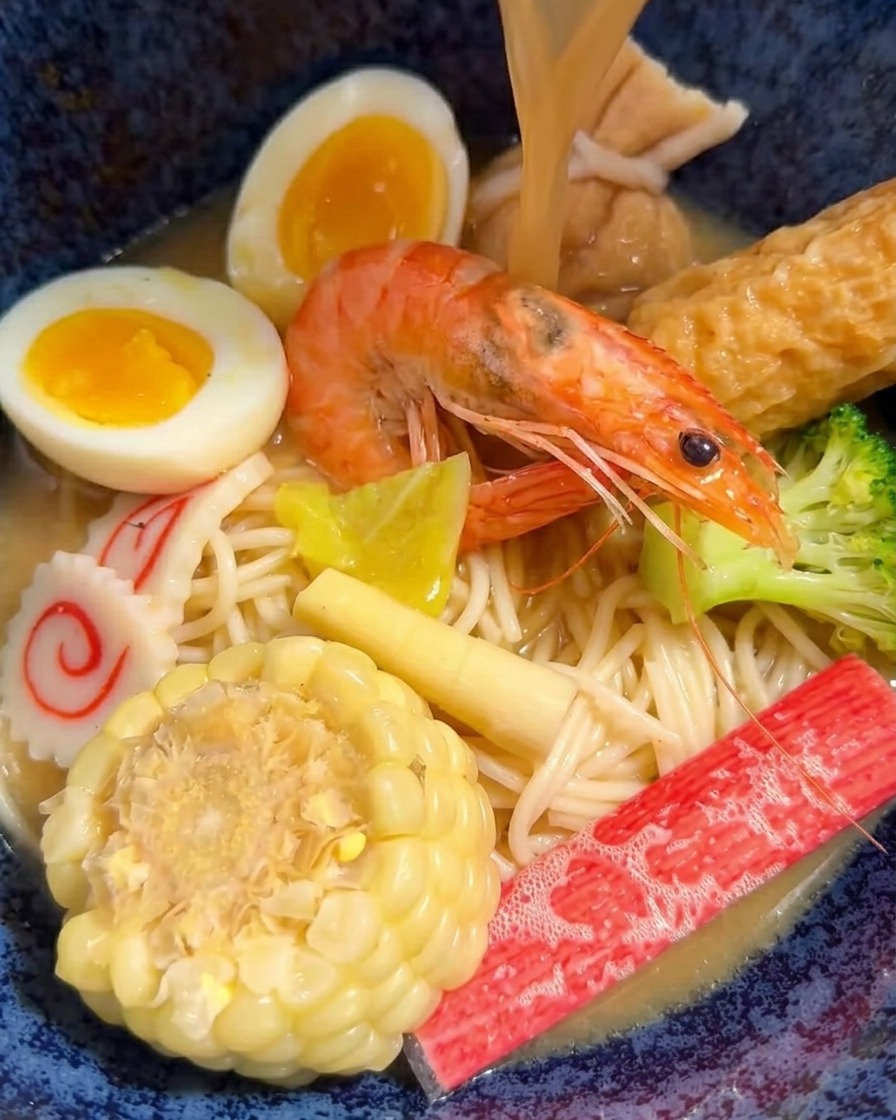 Hot miso broth being poured from a ladle into a bowl of ramen filled with various meats and vegetables.