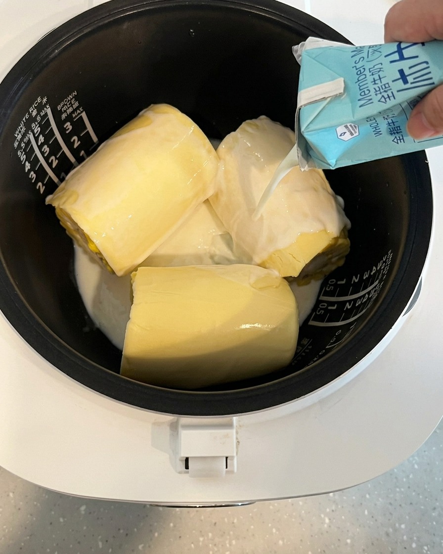 Milk being poured from a carton into a rice cooker pot containing corn and cheese.