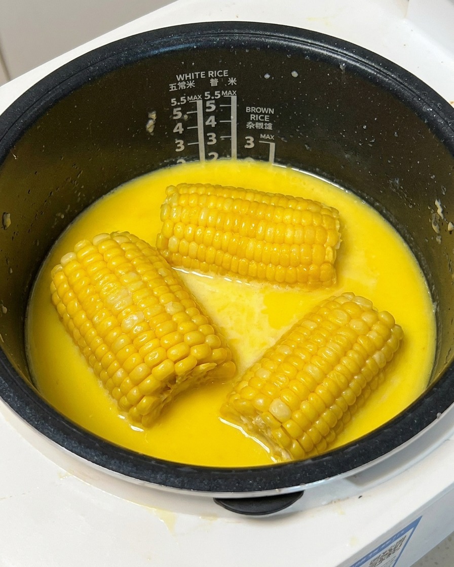 A cooked piece of yellow corn being lifted from a milky broth in a rice cooker using a wooden skewer.