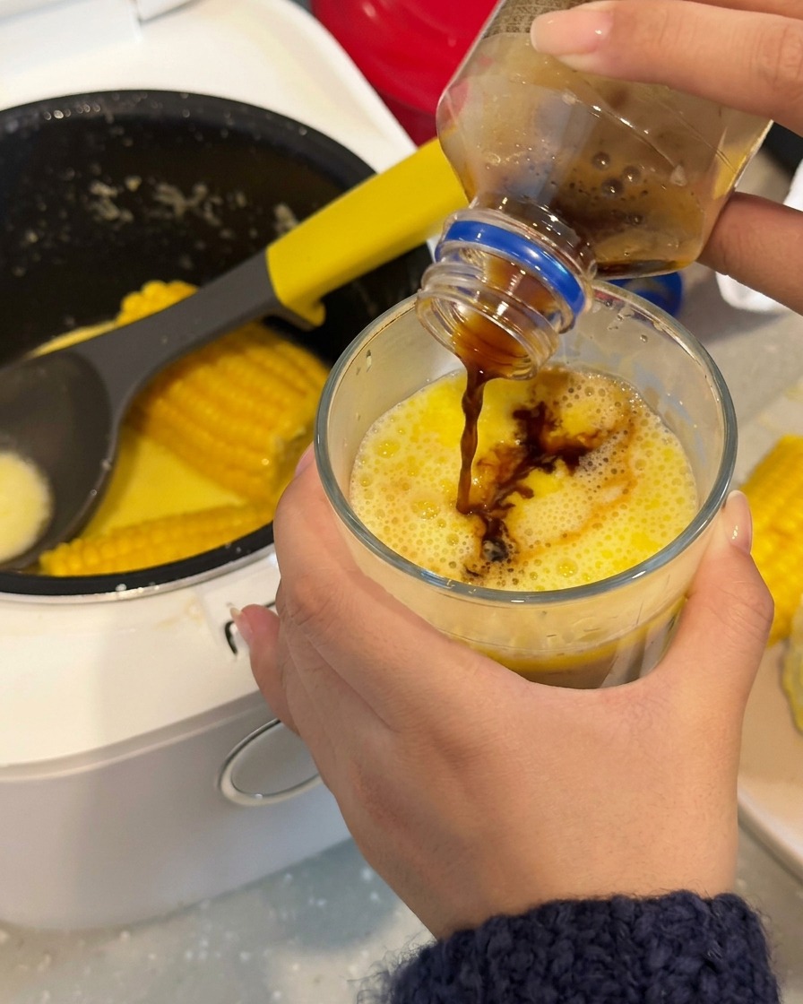 Dark coffee being poured into a glass containing a bright yellow, milky corn-infused broth.