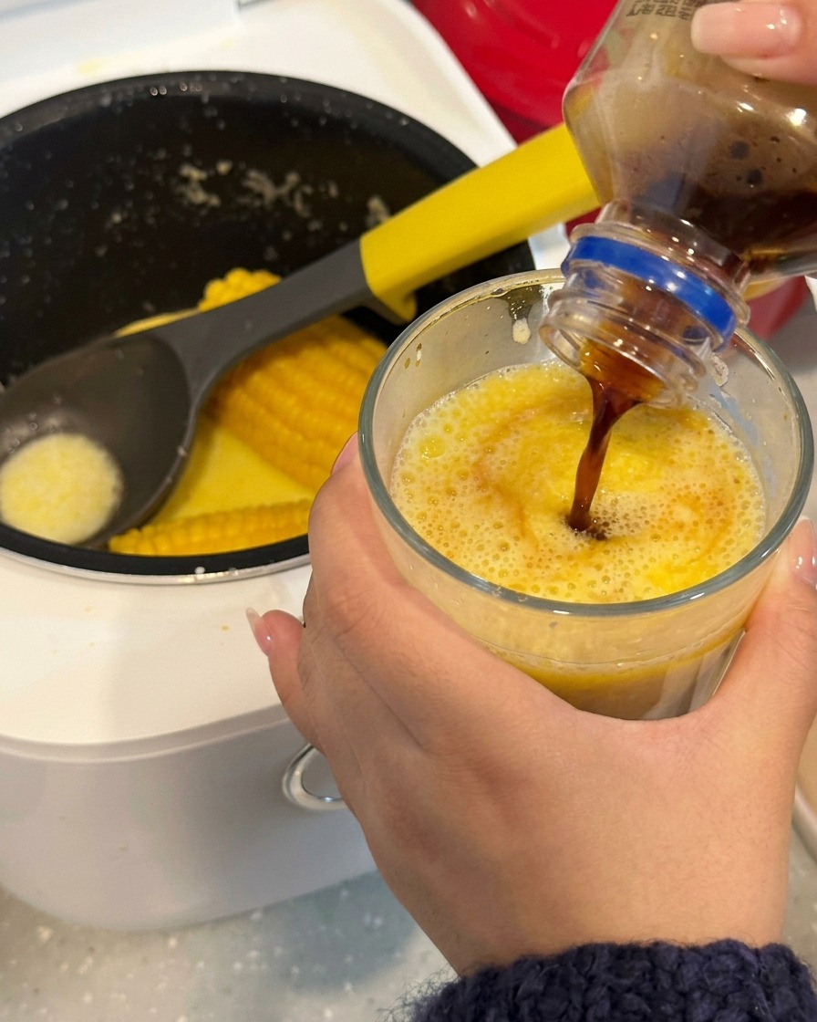 A close-up view of a wooden stick stirring a coffee and corn broth mixture in a clear glass.