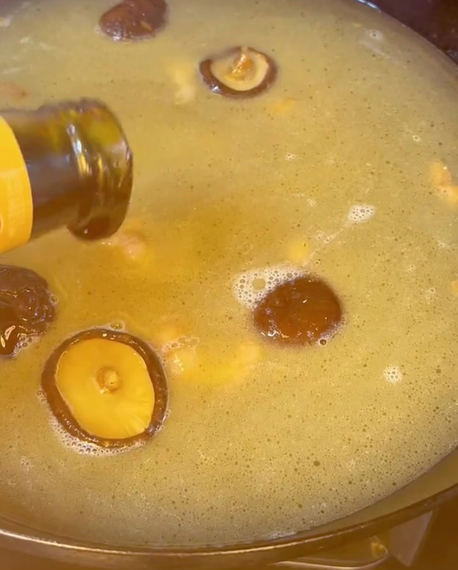 Oyster sauce being poured from a bottle into a boiling, golden broth filled with shiitake mushrooms.