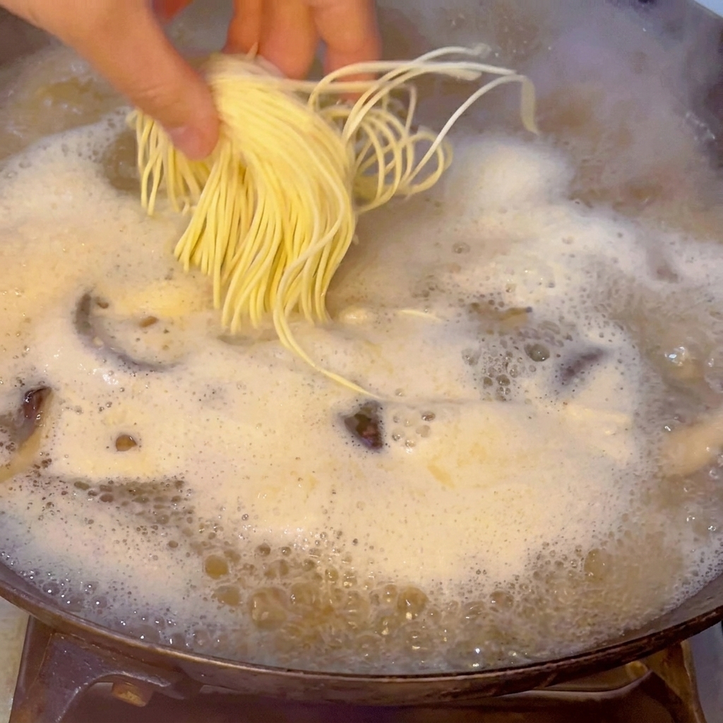 A hand carefully dropping a bundle of fresh, yellow ramen noodles into the boiling seafood broth.