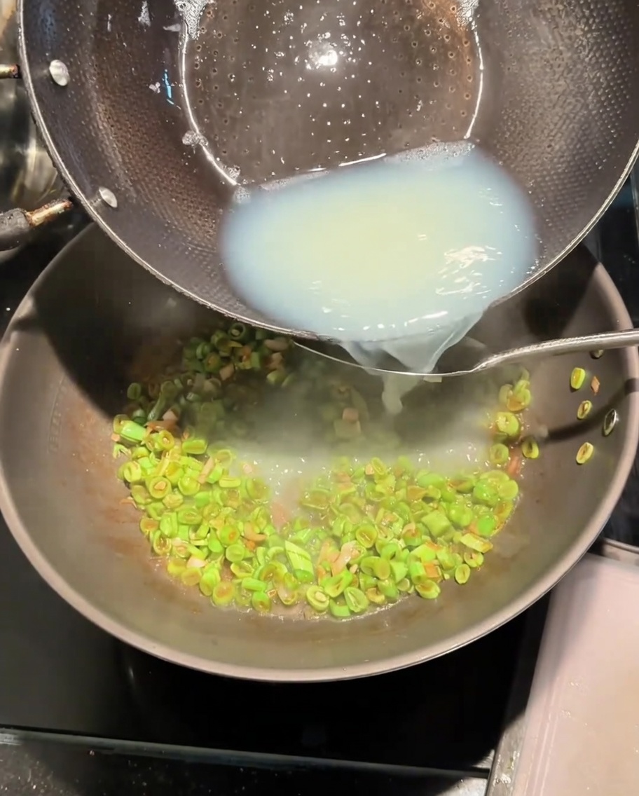 Pouring white clam broth from a pot into the wok with stir-fried beans.
