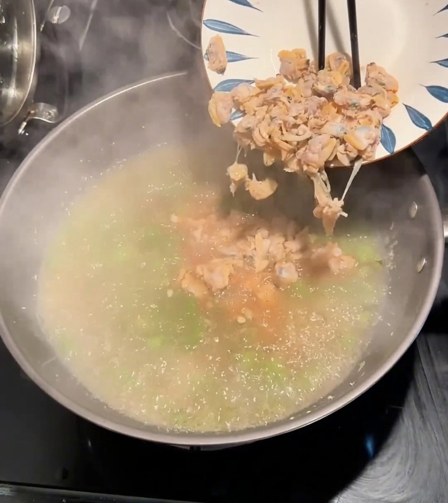 Clam meat being dropped from a plate into a pot of boiling seasoned broth with green vegetables.