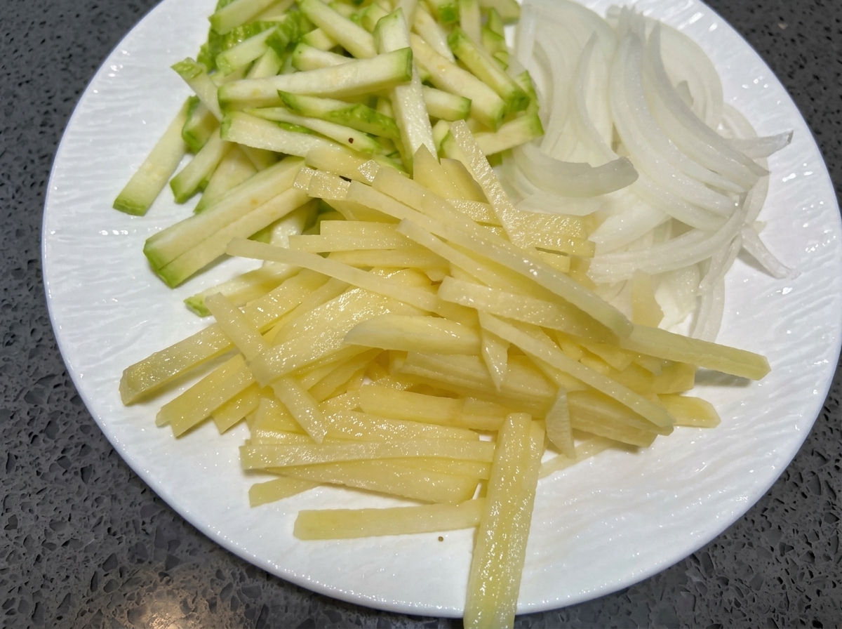 Thinly sliced strips of zucchini, onion, and potato resting on a white plate.
