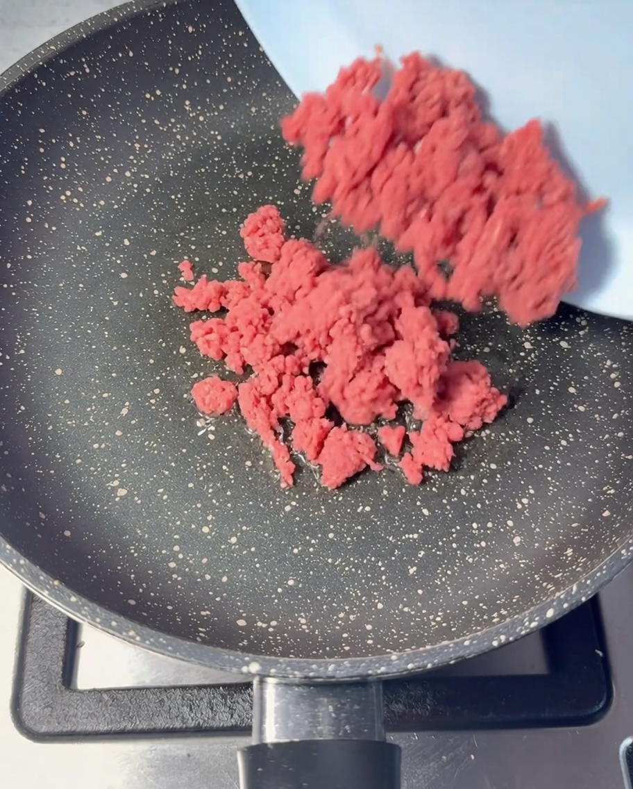 Raw pink minced beef being added to an oiled frying pan.
