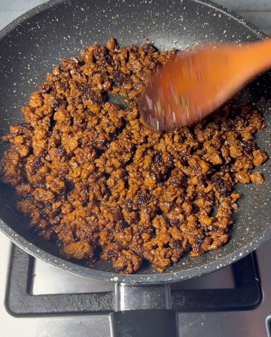 Minced beef being stir-fried in a speckled pan with a wooden spoon until fully browned.