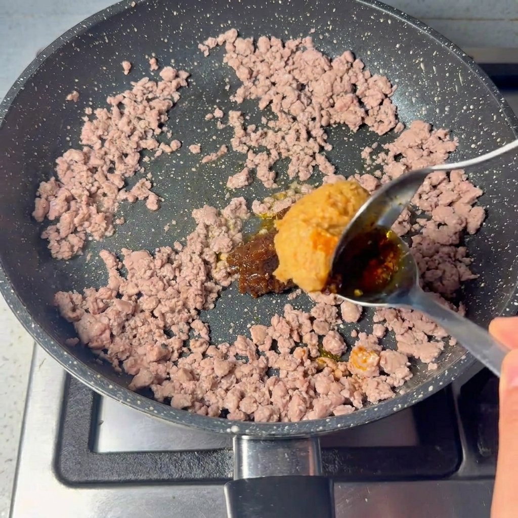 A large spoonful of thick brown paste being stirred into the cooked minced beef.