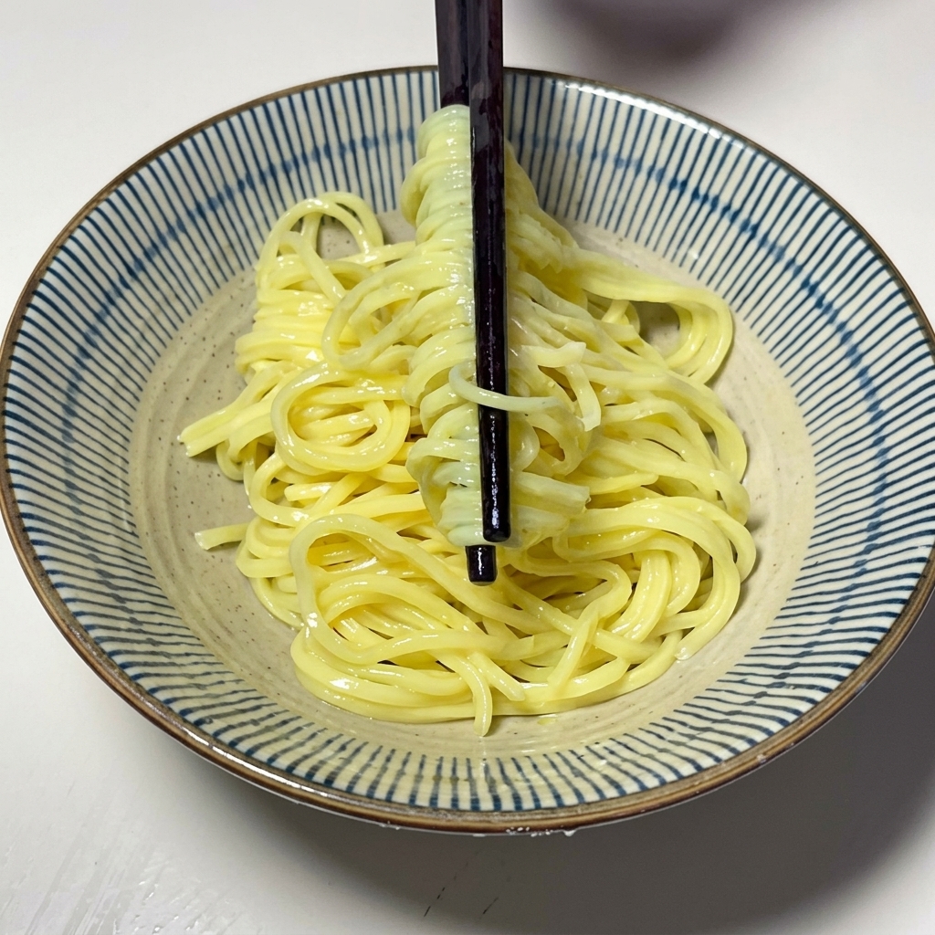 Cooked yellow noodles being lifted into a ceramic bowl with chopsticks.