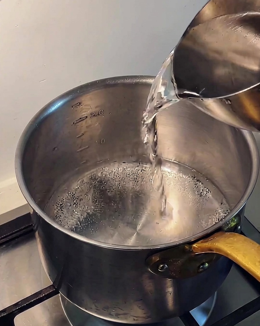 Pouring clear cold water from a metal pitcher into a stainless steel pot on a stove.