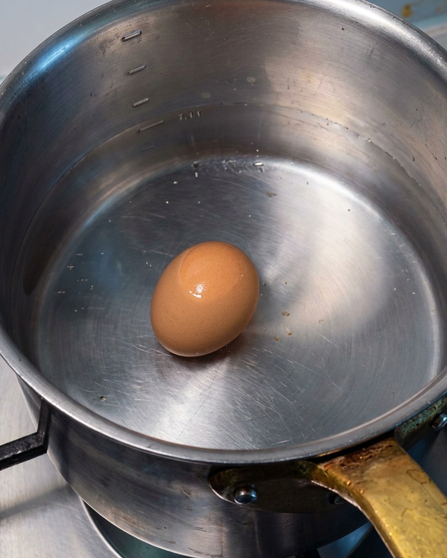 A whole brown egg resting at the bottom of a metal pot filled with hot water.