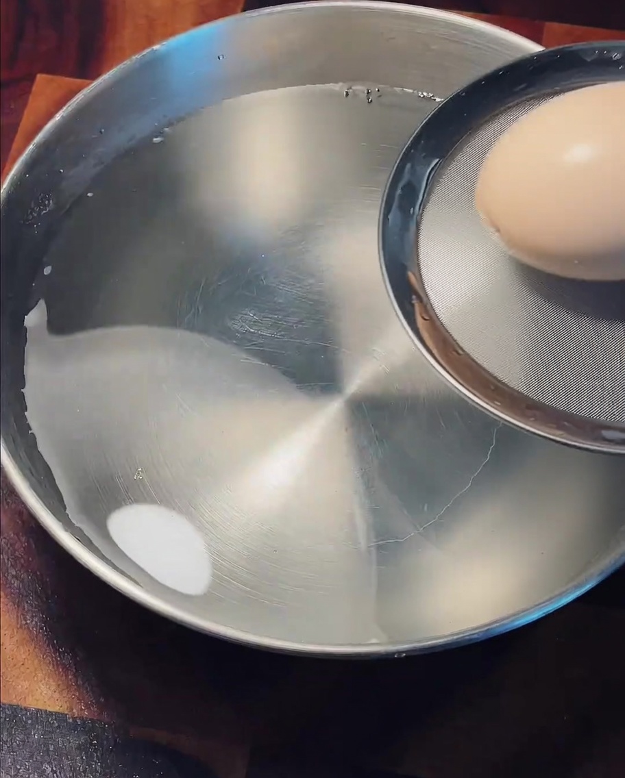 Lifting a cooked brown egg out of hot water using a wire mesh skimmer to transfer it into a cold water bath.