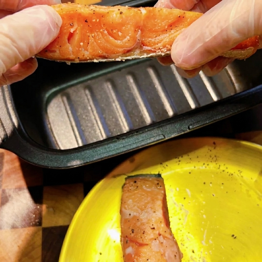 Gloved hands holding a seasoned raw salmon fillet over a dark rectangular microwave searing pan.