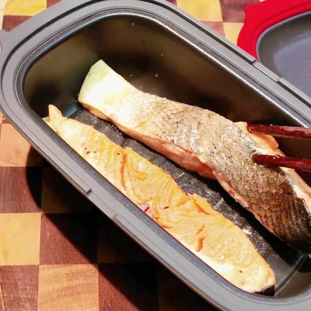 Chopsticks flipping a partially cooked salmon fillet inside a dark microwave cooking pan.