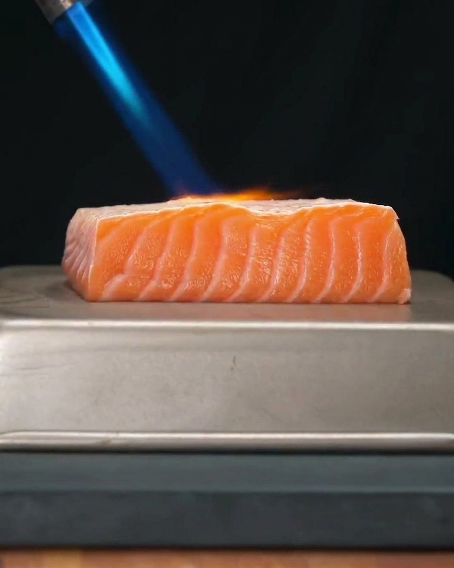 A thick, fresh salmon fillet being seared with a bright blue flame from a handheld kitchen torch.