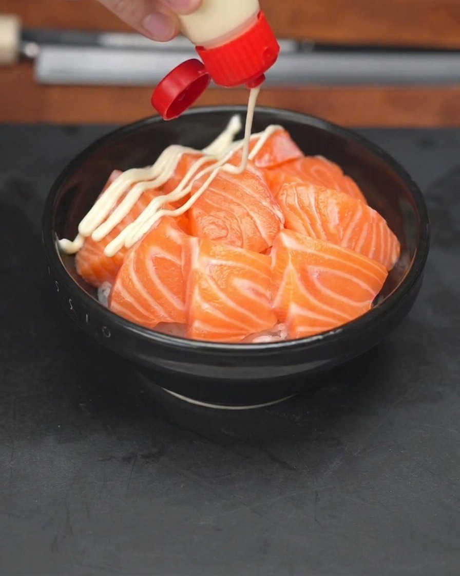 A hand squeezing a bottle of mayonnaise in a zig-zag pattern over a bowl of rice topped with salmon slices.