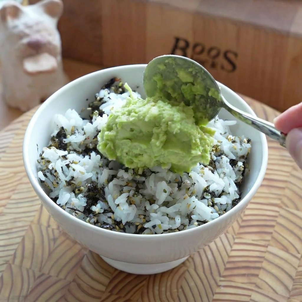 A layer of mashed avocado being spread smoothly over the rice base in a serving bowl.