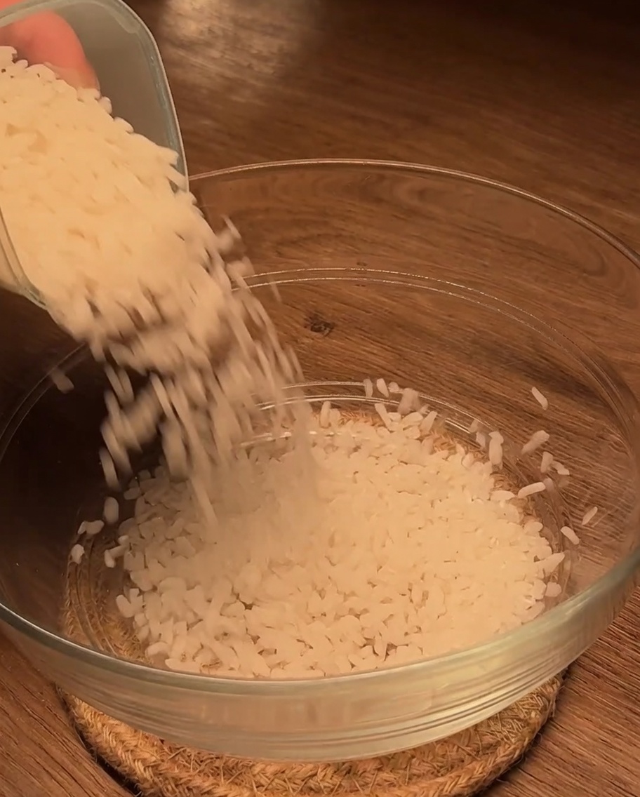 Uncooked white rice being poured into a clean glass bowl on a wooden surface.