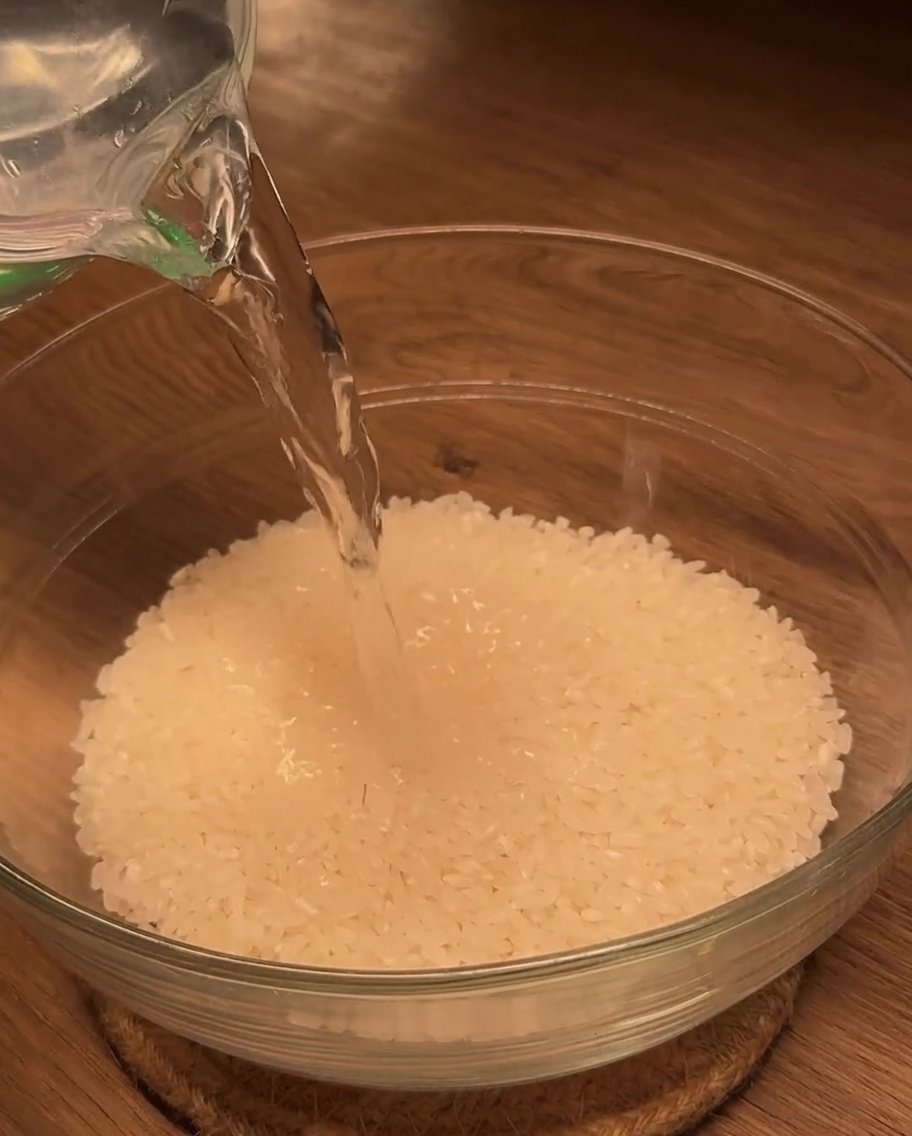 Water being poured from a measuring cup into a glass bowl filled with white rice.