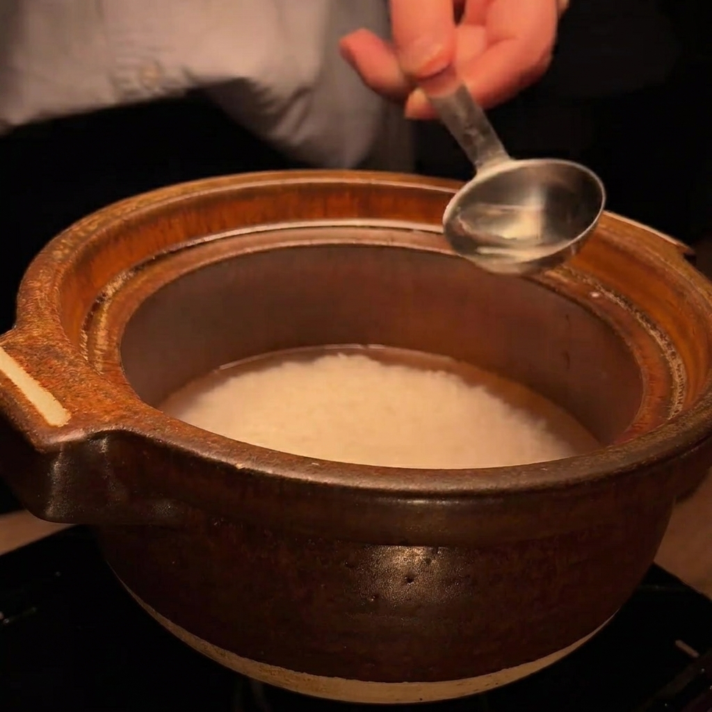 A metal spoon pouring a small amount of clear liquid, sake, into the clay pot with rice.