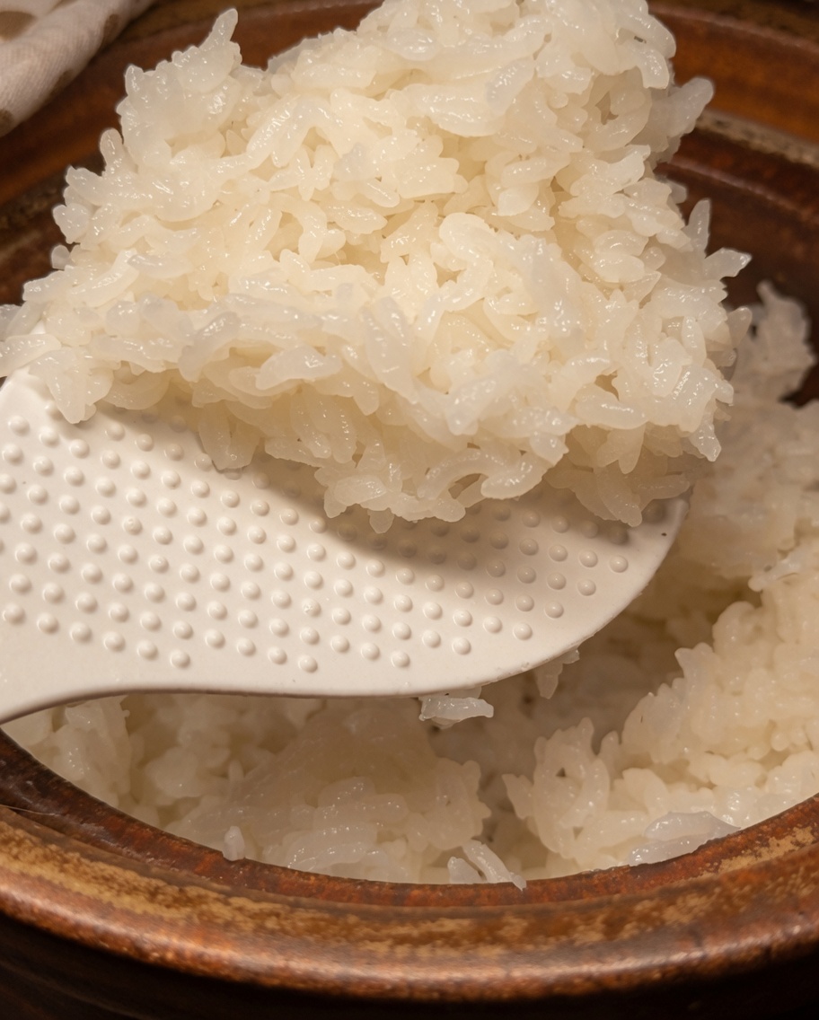 A close-up view of cooked white rice in a clay pot being gently mixed with seasoning using a white rice paddle.