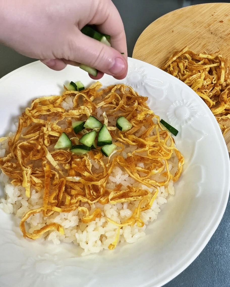 Diced green cucumber being sprinkled over the colorful layer of vinegared rice and egg strips.