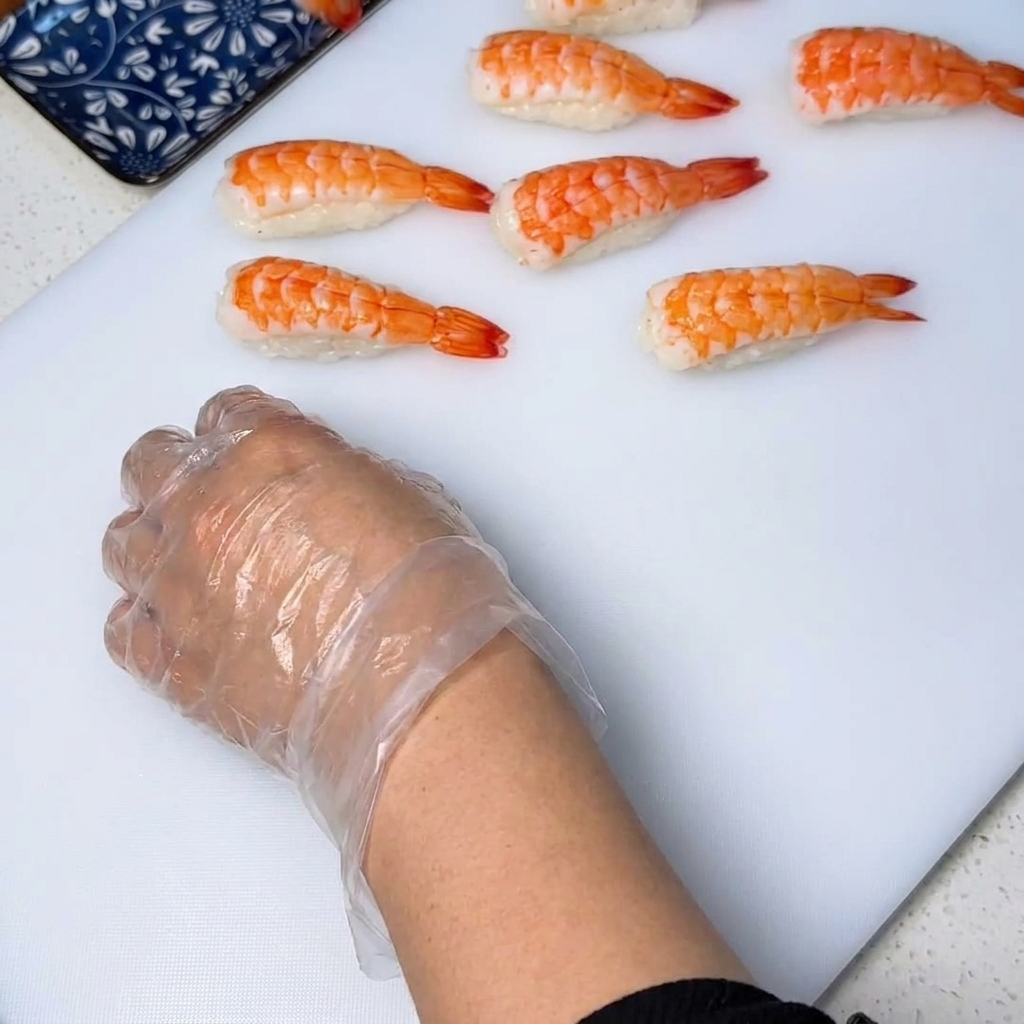 A close-up of a cooked shrimp being placed onto a small rice ball, with gloved hands pressing down to shape it into a neat piece of shrimp sushi.