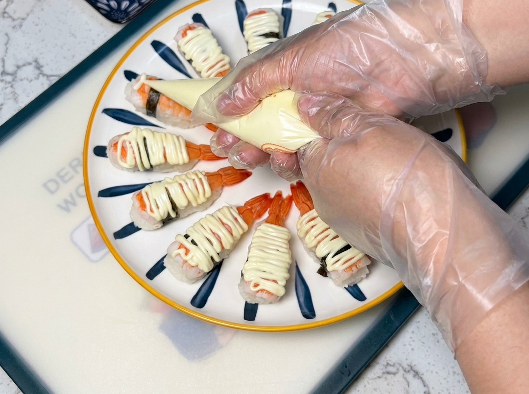 A close-up view of a piping bag squeezing mayonnaise over shrimp sushi arranged on a plate.