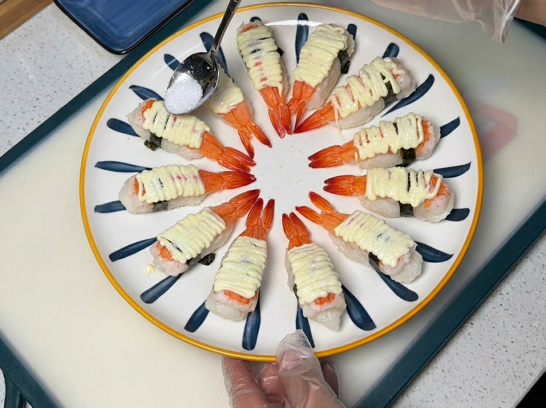 A spoon sprinkling sugar over mayonnaise-topped shrimp sushi on a decorative plate.