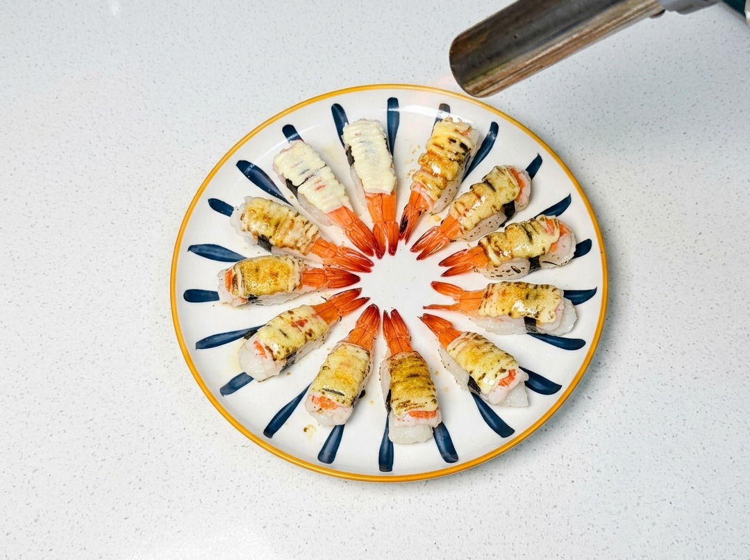 A culinary torch browning the sugar and mayonnaise topping on shrimp sushi pieces on a plate.