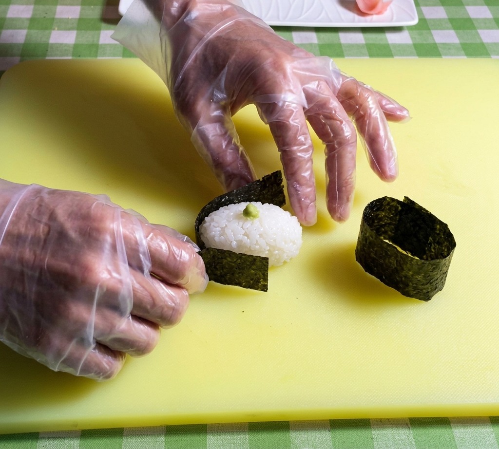 Hands placing a strip of dark nori seaweed around the edge of an oval-shaped sushi rice base.