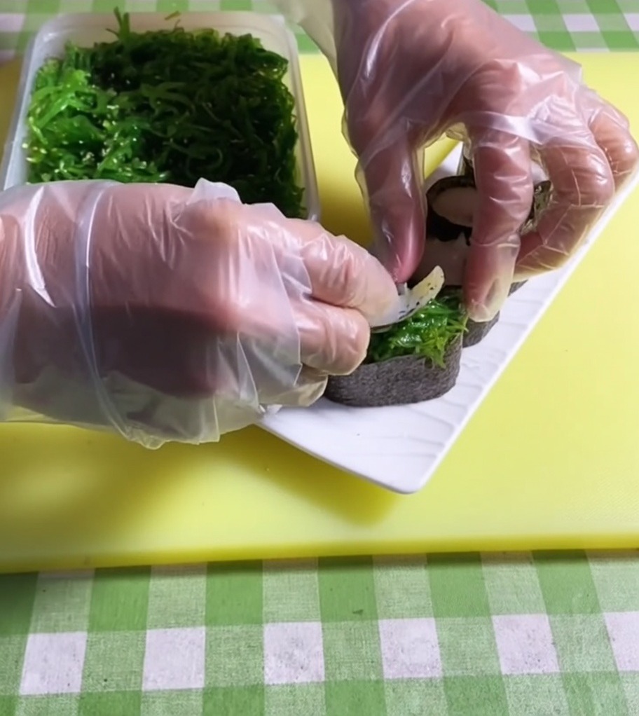 Tweezers placing a vibrant green portion of seaweed salad onto the top of a sushi base.