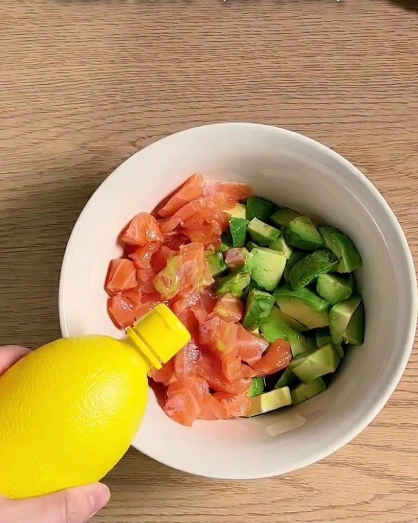 A bowl filled with cubed salmon and avocado being seasoned and mixed with various ingredients.