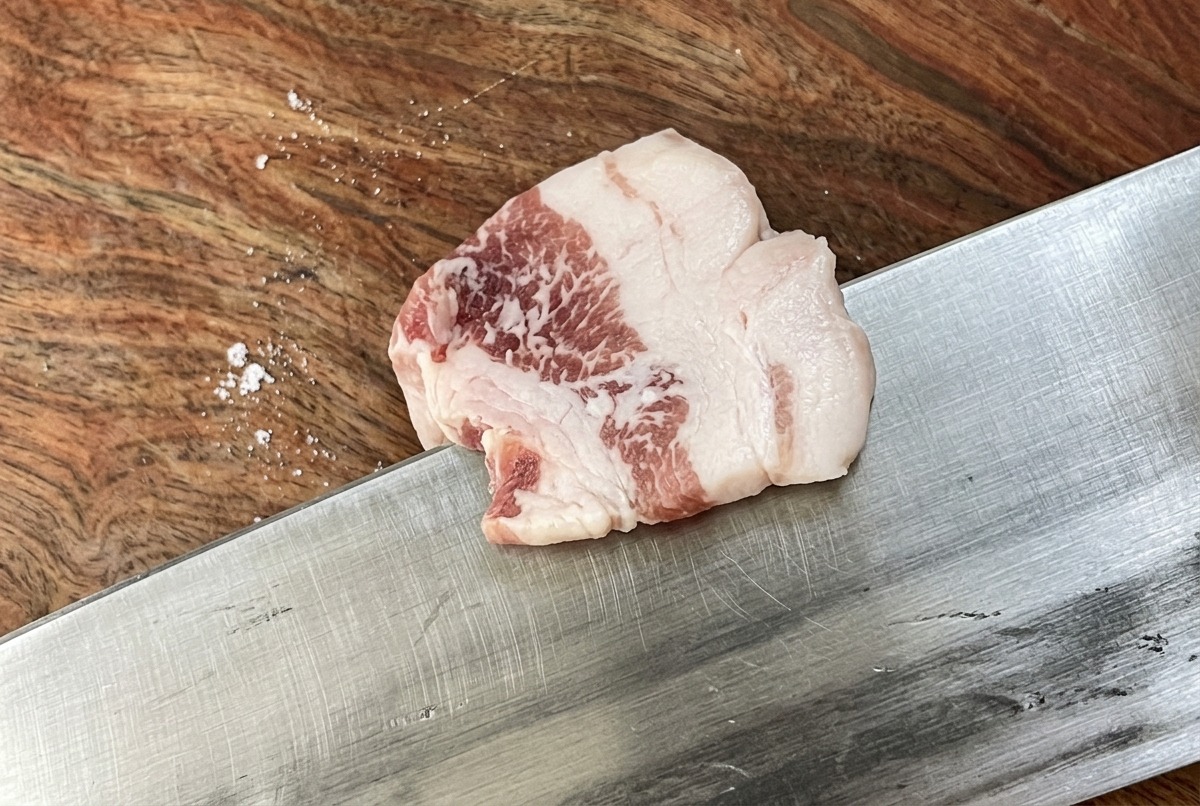 A sharp knife cutting a slab of raw pork belly into thin, uniform strips on a wooden cutting board.