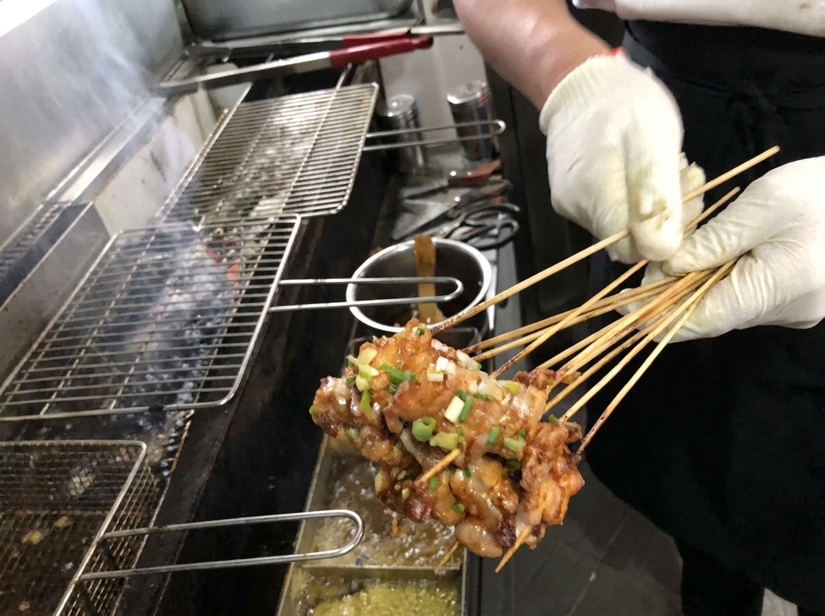 Close-up of a brush applying a rich, brown sauce onto golden-grilled pork belly skewers on a charcoal grill.