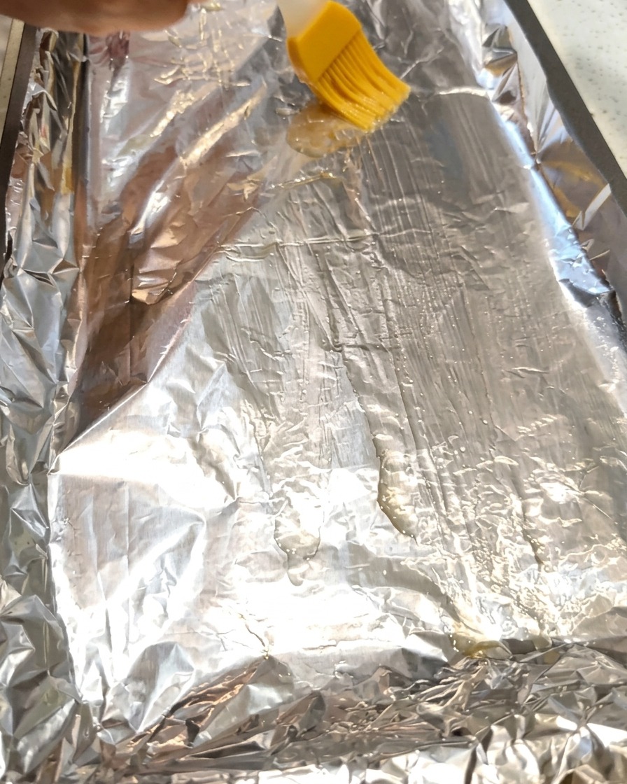 A brush spreading cooking oil onto a sheet of aluminum foil on a baking tray.