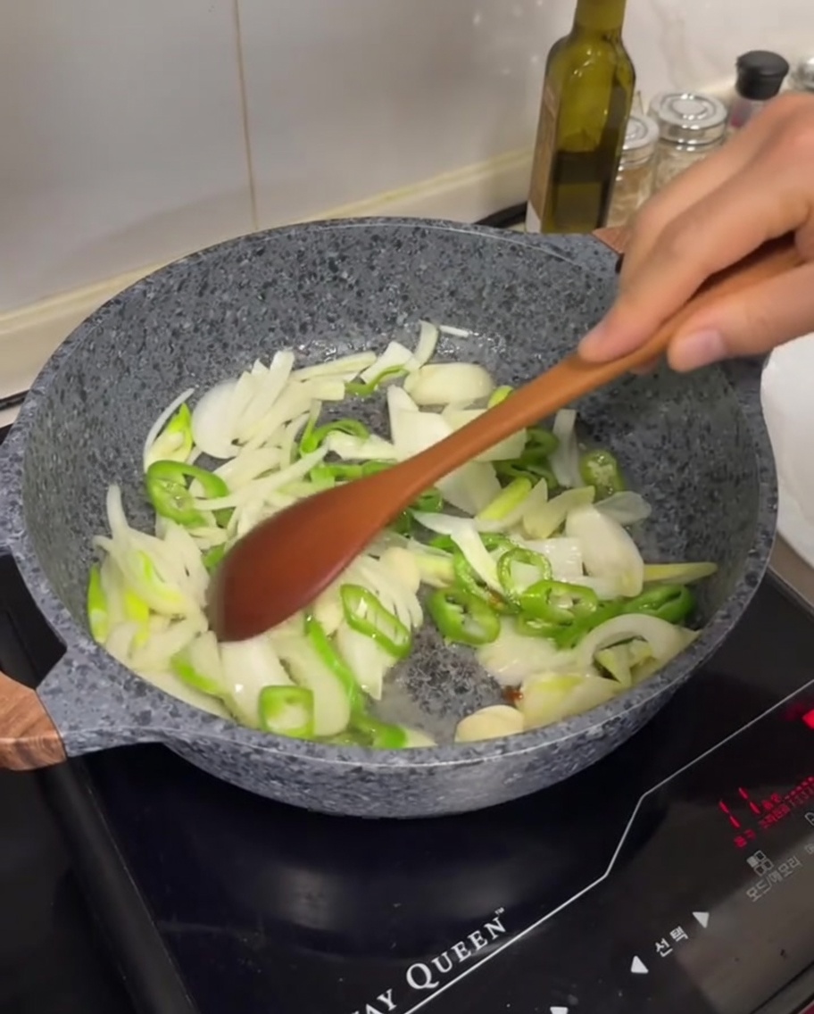 Sliced white onions, green chilies, and garlic cloves being stir-fried in a pan with rendered fat.