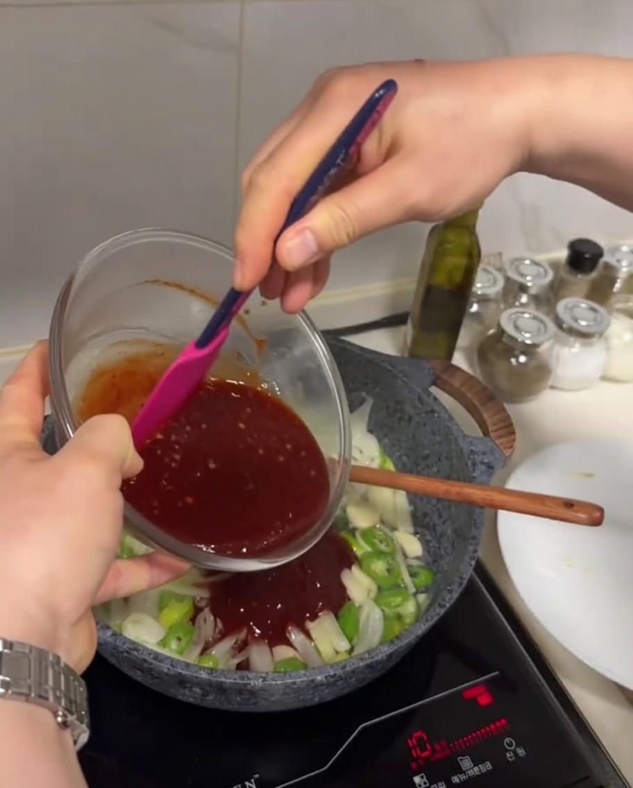 Dark red sauce being poured from a glass bowl into a pan containing sautéed vegetables.
