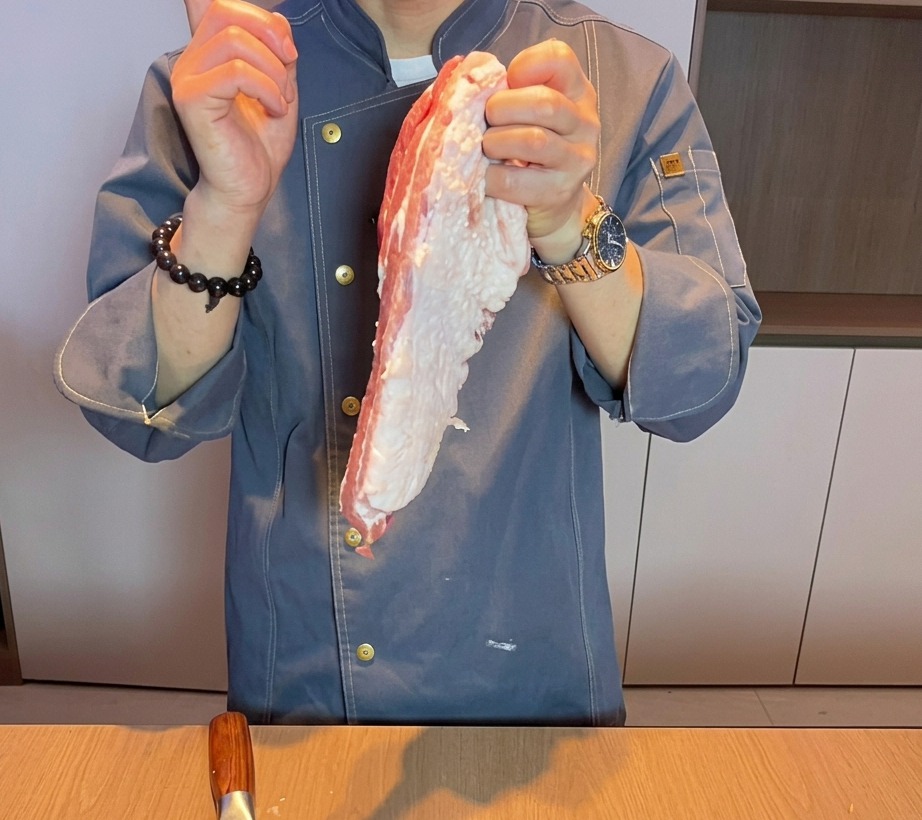 A close-up view of a chefs hand cutting fresh, raw pork belly into thick chunks on a wooden cutting board.