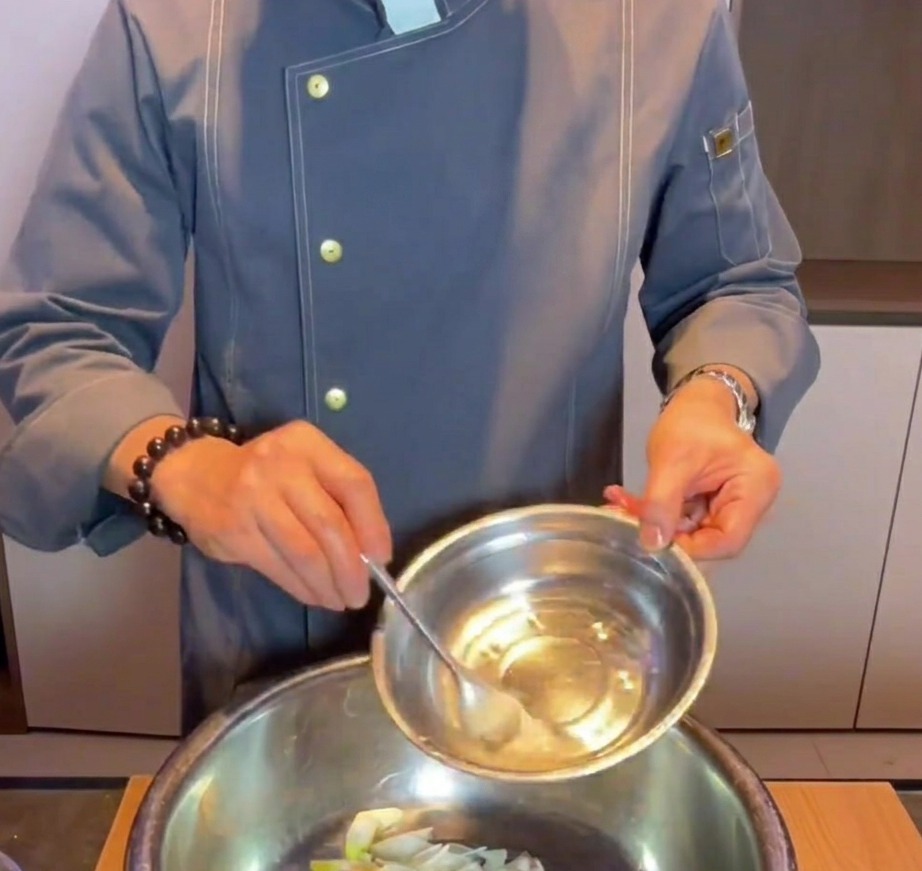 Hand pouring baking soda water into a metal mixing bowl with raw pork belly, ginger, and green onion slices.